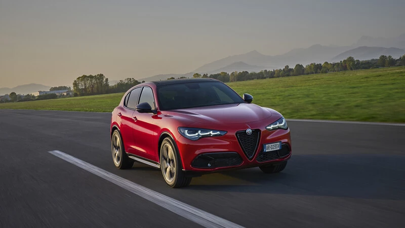 Červené Alfa Romeo Stelvio v jazde po ceste s horami a zelenými lúkami v pozadí