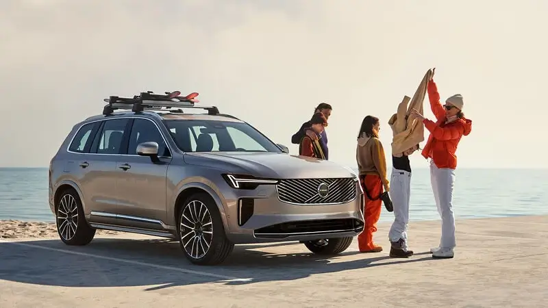 Sivé Volvo XC90 so strešným nosičom a lyžiarskou výbavou na pobreží – rodinné 7-miestne prémiové SUV pripravené na víkendové výlety