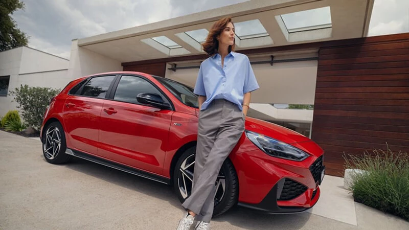 Žena opretá o červený Hyundai i30 N Line pred moderným rodinným domom