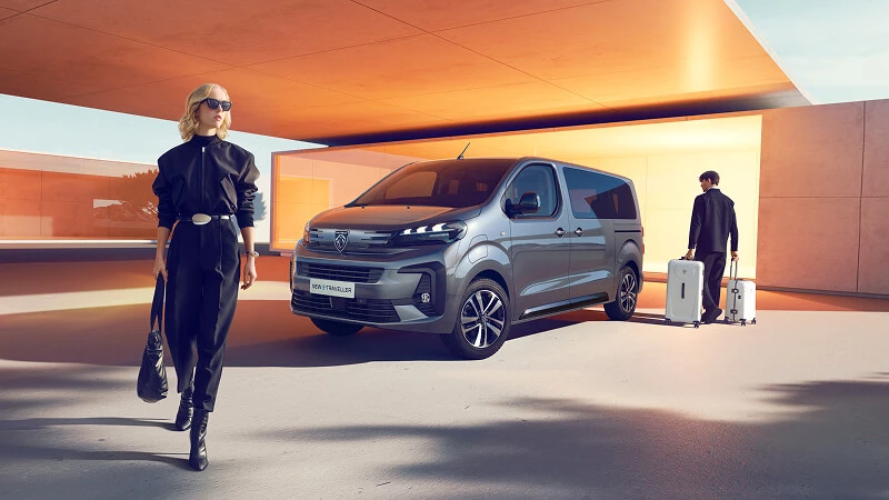 Cestujúci s kuframi pri Peugeote Traveller – sivý minibus na prepravu osôb pred moderným hotelom